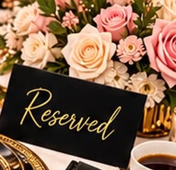 A black Reserved place card in front of a bouquet of pink and cream roses, beside a gold charger plate and a cup of coffee.